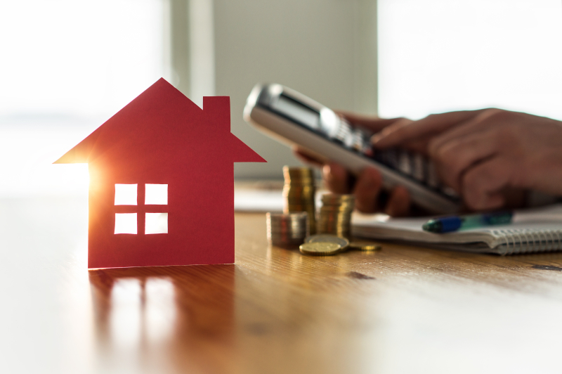 les taux de prêt immobilier 2022 - A côté d’une petite maison et de quelques pièces de monnaie en colonne, une main tient une calculatrice pour évaluer le coût total d’un projet immobilier
