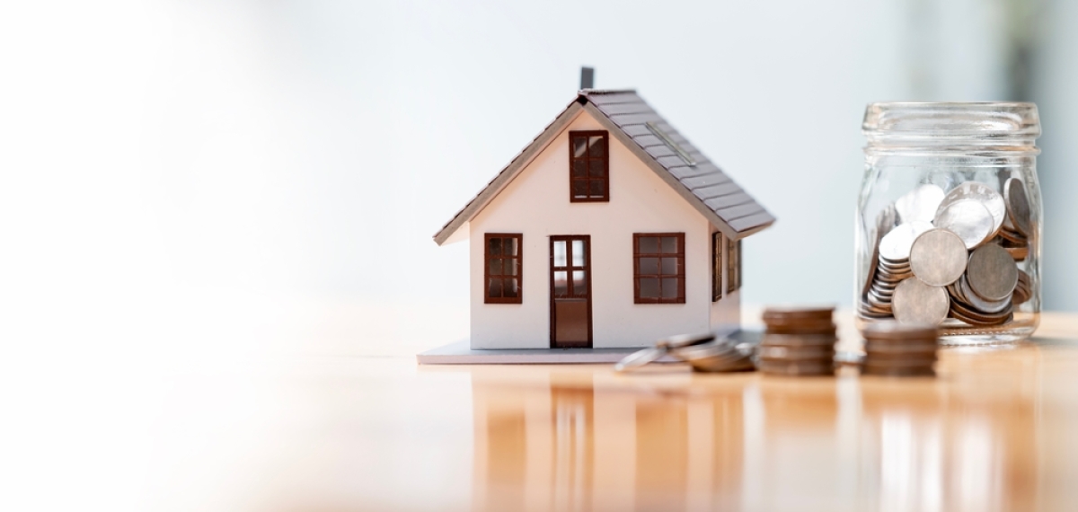 Normes PMR logement neuf — Une maison miniature posée sur un bureau à côté d’un pot rempli de pièces de monnaie