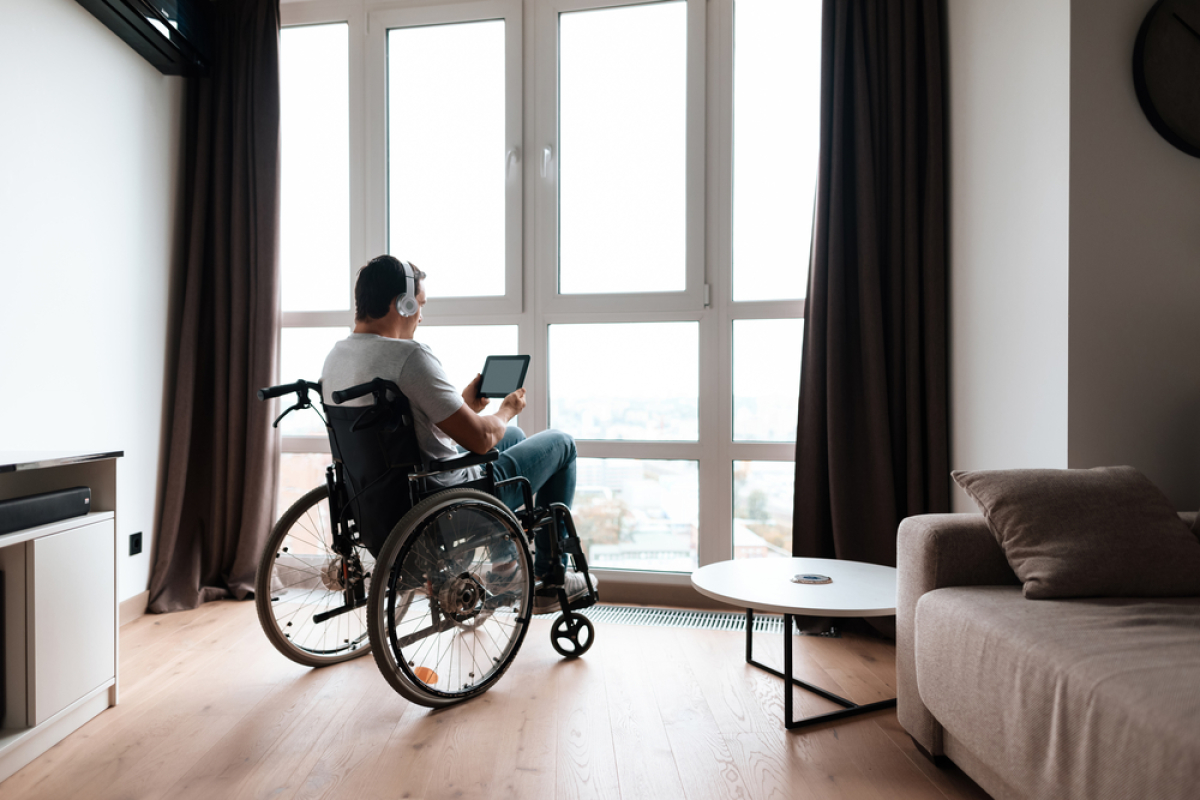Normes PMR logement neuf — Une personne en fauteuil roulant regardant sa tablette à proximité d’une baie vitrée